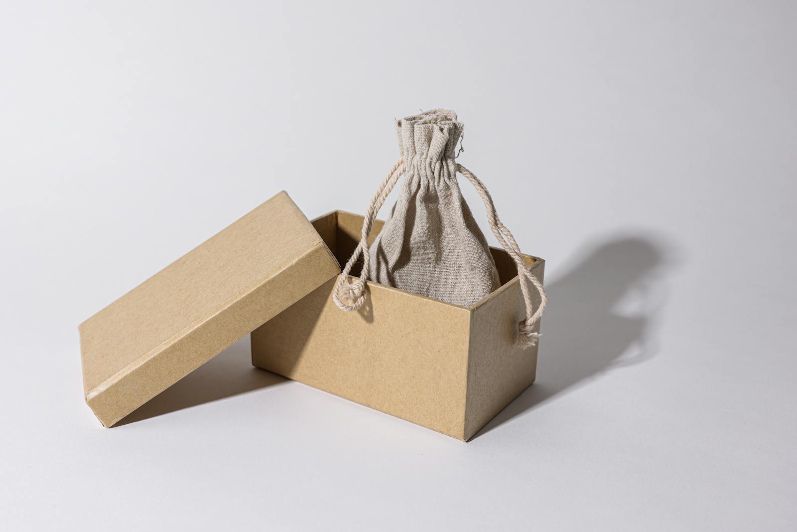A cardboard box with a linen pouch inside, casting a shadow on a plain background.